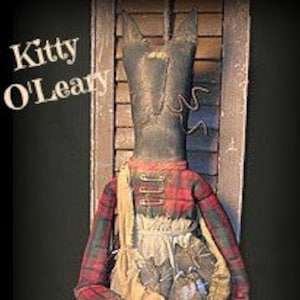 Könnte beinhalten: Eine handgemachte Stoffpuppe mit schwarzem Gesicht, rot-schwarzem kariertem Hemd und einer braunen Tasche. Die Puppe heißt "Kitty O'Leary".
