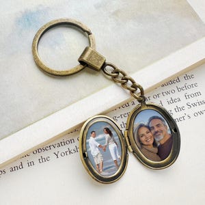 May include: A bronze-colored metal keychain with an oval locket. The locket opens to reveal two small photographs of couples. The keychain is on a light-colored surface with text.