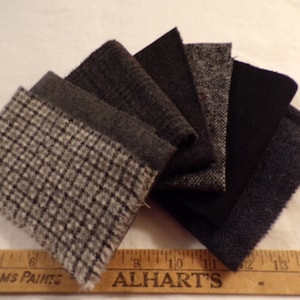 May include: A collection of seven rectangular fabric swatches in various shades of gray, black, and navy blue, featuring textures like houndstooth, tweed, and solid wool, positioned above a wooden ruler measuring from 4 to 14 inches.