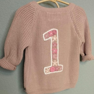 以下が含まれることがあります： ピンクのニットセーターで、背中に大きな刺繍の数字1が施されています。数字1はピンクと白の花で飾られています。