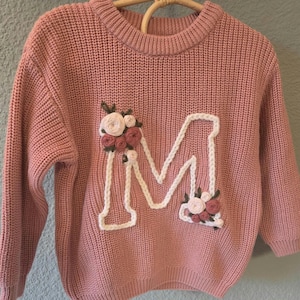 May include: A pale pink knitted sweater featuring a large white embroidered letter "M", decorated with floral details in pink, white, and green. The sweater is hanging on a wooden clothes hanger.