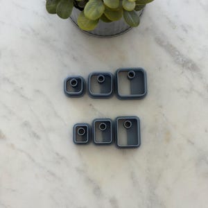 May include: A set of six gray plastic clay cutters arranged in two rows. The top row features three square-shaped cutters of increasing size, while the bottom row contains three rectangular-shaped cutters of increasing size, each with a center hole.