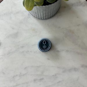 May include: A small, round, dark blue wax seal with a white snowflake design in the center. The seal is on a white marble surface, with a small metal pot of greenery in the background.