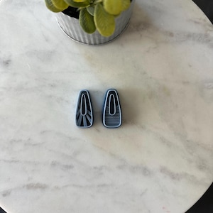 May include: Two blue and black hair clips with geometric designs. One clip has a striped pattern, and the other has a fan-like design. The clips are on a white marble surface, with a small potted plant in the background.
