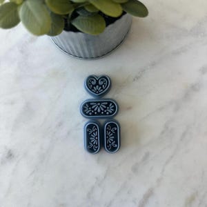 May include: A collection of three blue cookie cutters in heart, oval, and rectangular shapes, each with a floral design. The cutters are arranged vertically on a white marble surface, with a small potted plant in the background.