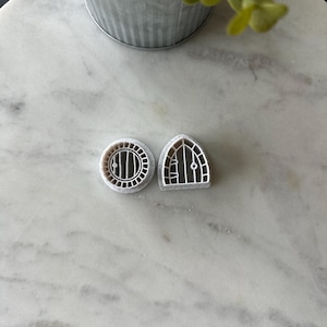 May include: Two white plastic cookie cutters, one shaped like a round door with a circular window and the other shaped like a pointed arched door with a rectangular window.