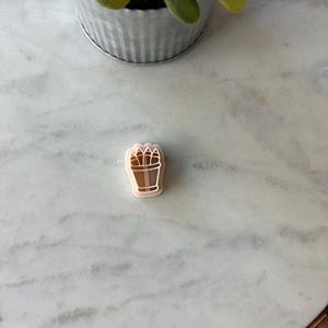 May include: A light pink cookie cutter in the shape of a potted plant. The plant has a brown pot and brown leaves. The cookie cutter is on a white marble surface.