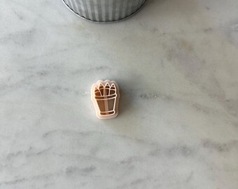 May include: A light pink cookie cutter in the shape of a potted plant. The plant has a brown pot and brown leaves. The cookie cutter is on a white marble surface.