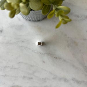 May include: A white ceramic ring dish with a rounded shape and a small opening in the center. The dish is sitting on a white marble surface.