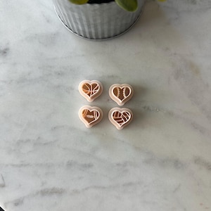 May include: Four heart-shaped cookie cutters with sports ball designs: basketball, baseball, football, and volleyball. The cutters are light pink and sit on a white marble surface. A small metal pot with a green plant is in the background.