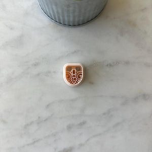 May include: A small, light-colored cookie cutter with a decorative design. The cutter is square with rounded corners and features an intricate pattern within its center. The cutter is on a white marble surface, with a plant in a metal pot in the background.