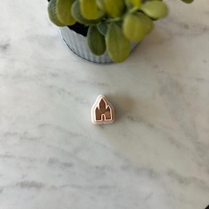 May include: A small, light pink and brown castle-shaped cookie cutter. The cutter is made of plastic and has a detailed design. The castle is centered on a white marble surface, with a small potted plant in the background.