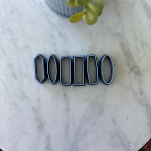 May include: A set of six blue plastic cookie cutters in various shapes, including a rectangle, oval, and a pointed shape. The cutters are on a white marble surface.