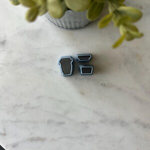 May include: Three silicone cookie cutters in the shape of coffee cups. The cutters are gray with black outlines.