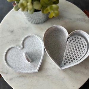 May include: A white heart-shaped trinket box with a removable lid. The lid has a small handle. The box has a patterned interior with small heart cutouts.