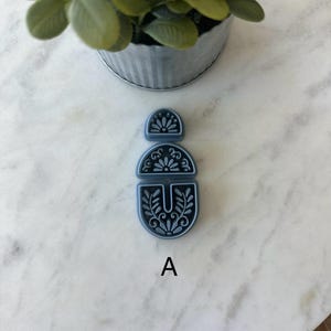 Op de afbeelding: Een driedelige, donkerblauwe oorbelcomponentenset met een bloemmotief. De componenten zijn verticaal gerangschikt, met de letter "A" onder het onderste stuk. De set ligt op een wit marmeren oppervlak.