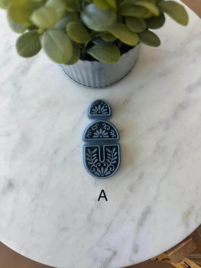 Op de afbeelding: Een driedelige, donkerblauwe oorbelcomponentenset met een bloemmotief. De componenten zijn verticaal gerangschikt, met de letter "A" onder het onderste stuk. De set ligt op een wit marmeren oppervlak.