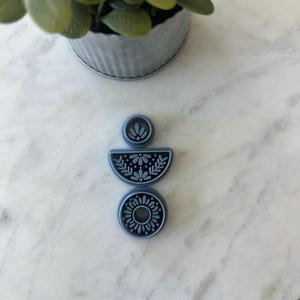 Op de afbeelding: Een driedelige, blauwgrijze polymeerklei hanger met bloemen- en bladerpatronen. De hanger bestaat uit een kleine cirkel, een halve cirkel en een volledige cirkel, elk met een gedetailleerd ontwerp. De hanger wordt gepresenteerd op een wit marmeren oppervlak.