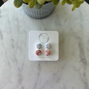 May include: Four floral stud earrings on a white card. The earrings feature red, blue, and white floral patterns. The card has the text "Handcrafted Jewellery" and a logo. The earrings are displayed on a marble surface.