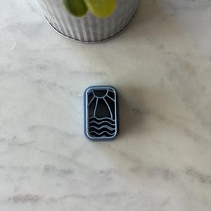May include: A rectangular, blue and white cookie cutter featuring a sun and wave design. The cutter is made of plastic and is designed for use with cookie dough or fondant. The design includes a sun with rays and three wavy lines.