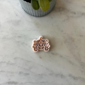 May include: A light brown, plastic cookie cutter in the shape of a fairytale carriage. The carriage has detailed wheels, windows, and a decorative top. The cutter is designed for creating cookies or other treats with a whimsical theme.