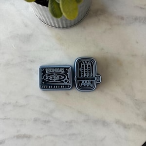 May include: Two blue sardine-themed cookie cutters. One is rectangular with the word "Sardines" and a fish illustration. The other is an open can shape with sardines inside. The cutters are on a marble surface.