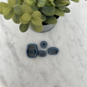 May include: Five blue plastic cookie cutters in the shape of a jar, a small circle, a rectangle, and two different sized pie crusts. The cutters are on a white marble surface.
