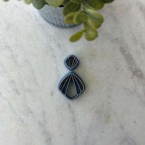May include: A light blue polymer clay cutter in a fan shape, with a smaller, diamond-shaped cutter above it. The cutters are on a white and gray marble surface, with a small metal bucket of green leaves in the background.
