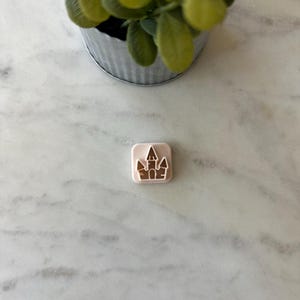 May include: A small, square, light pink object with a white castle design. The castle features multiple towers and a detailed facade. The object is set on a white and gray marble surface, with a small metal pot of green plants in the background.