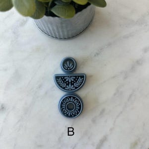 Op de afbeelding: Een set van drie blauwe polymeerklei uitsteekvormen met bloemmotieven. De uitsteekvormen hebben de vorm van een cirkel, een halve cirkel en een kleine cirkel. De letter "B" is wit gedrukt.