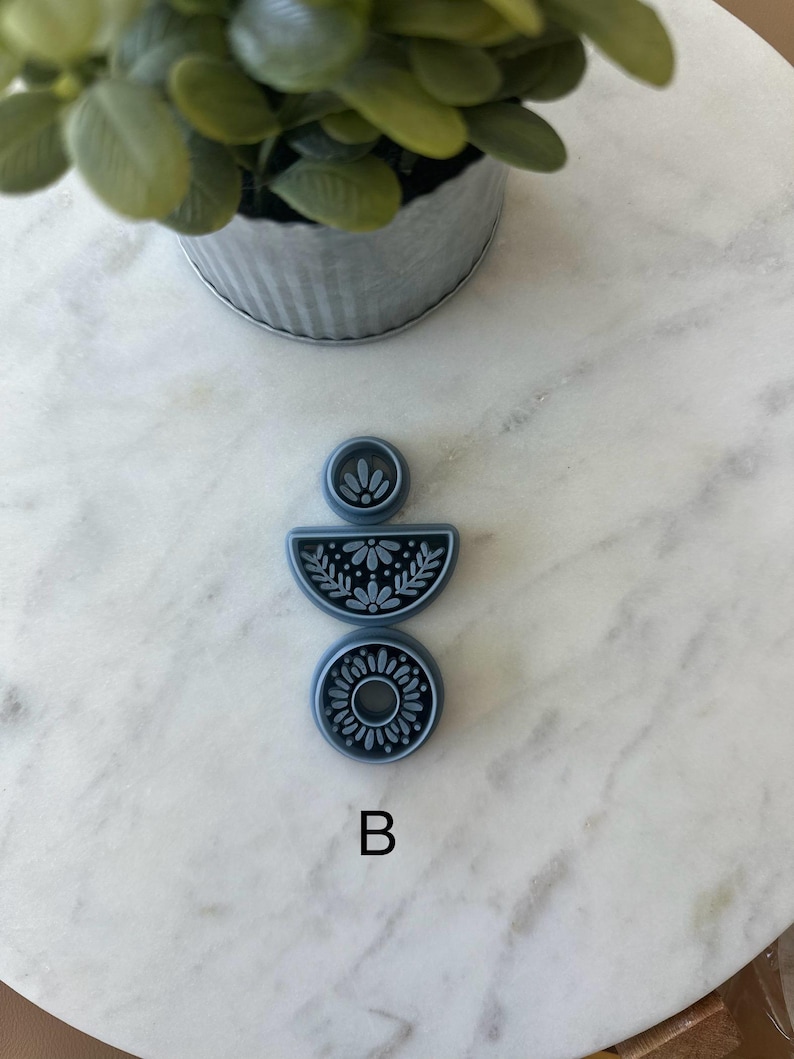 Op de afbeelding: Een set van drie blauwe polymeerklei uitsteekvormen met bloemmotieven. De uitsteekvormen hebben de vorm van een cirkel, een halve cirkel en een kleine cirkel. De letter "B" is wit gedrukt.
