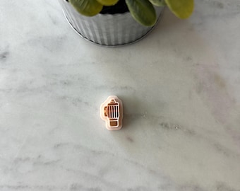 May include: A small, light pink fire hydrant-shaped cookie cutter with brown details. The cookie cutter is on a white marble surface, with a small potted plant in the background.