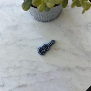 May include: A small, dark blue violin-shaped object rests on a white marble surface. The object has a detailed outline, suggesting a cookie cutter or decorative element. A small, galvanized metal pot with green foliage is in the background.