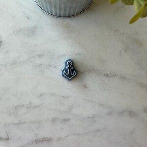 May include: A small blue ceramic anchor charm with a white outline. The anchor is on a white marble surface.