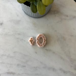May include: Two light pink cookie cutters, one in the shape of a frame and the other in the shape of a ghost. The frame cutter has an oval center with a decorative border. The ghost cutter is a simple outline. The cutters are on a white marble surface.