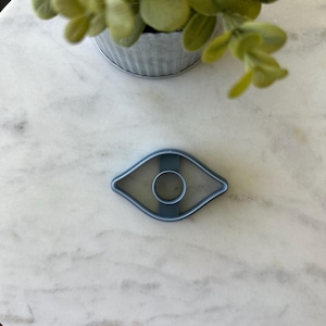 May include: A blue eye-shaped cookie cutter with a circular center, resting on a white marble surface. The cutter is made of plastic and has a smooth finish. A small metal pot with green leaves is in the background.
