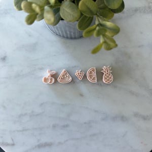 May include: A collection of five fruit-shaped cookie cutters: cherry, watermelon slice, grapes, orange slice, and pineapple. The cutters are light pink and arranged in a row on a marble surface. A small metal bucket with greenery is in the background.