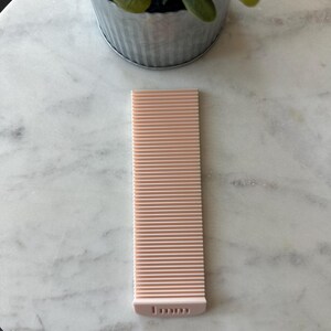 May include: A light pink, rectangular tool with parallel ridges, possibly a crafting or measuring tool. The tool has a rounded end with the text "1 mm" printed on it. The tool is lying on a marble surface, with a small potted plant in the background.