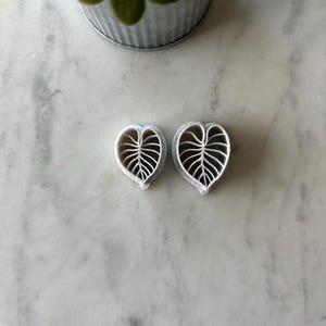 May include: Two white heart-shaped earrings with a leaf design. The earrings are on a white marble surface.
