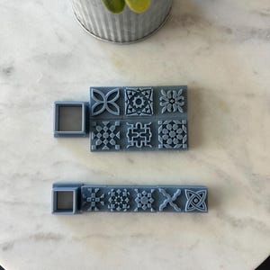 May include: A collection of gray square tile stamps with various geometric patterns. The stamps are arranged on a white and gray marble surface. A small square frame is also included.