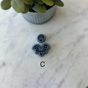 Op de afbeelding: Een kleine, blauwe bloemvormige polymeerklei-uitsteker met een bijpassende ronde bovenkant en de letter "C" in zwart. De uitsteker staat op een wit marmeren oppervlak, met een kleine plant in een pot op de achtergrond.