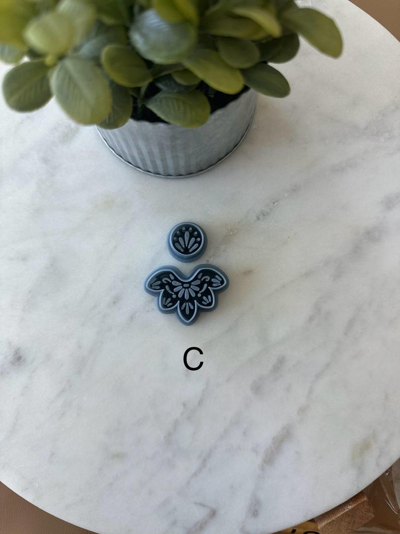 Op de afbeelding: Een kleine, blauwe bloemvormige polymeerklei-uitsteker met een bijpassende ronde bovenkant en de letter "C" in zwart. De uitsteker staat op een wit marmeren oppervlak, met een kleine plant in een pot op de achtergrond.