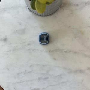 May include: A small, blue and white number zero cookie cutter. The cookie cutter is on a white marble surface. A small metal pot with green leaves is in the background.