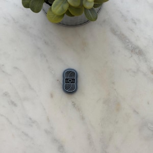 May include: A small, oval-shaped charm featuring a blue-grey background with a white line drawing of a ball of yarn. The design includes a horizontal band across the center of the yarn ball with a small, centered heart icon in white outline.