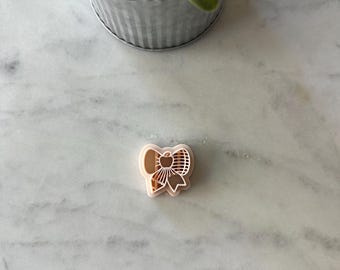 May include: A light pink bow-shaped cookie cutter with a detailed design. The cutter is made of plastic and is designed for creating bow-shaped cookies or fondant decorations. The cutter is on a marble surface.