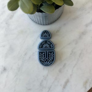 Op de afbeelding: Een driedelige, donkergrijze polymeerklei-uitsteker set. Elk stuk heeft een bloemenontwerp. De uitsteker set ligt op een wit marmeren oppervlak, met een kleine potplant op de achtergrond.