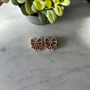 May include: Two pink plastic cookie cutters shaped like bows with a gold design. The cookie cutters are on a white marble surface.