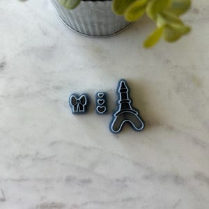 May include: A set of three blue plastic cookie cutters. The cutters are shaped like a bow, two hearts, and the Eiffel Tower.