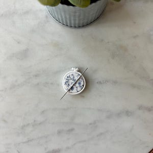 May include: A small, white embroidery hoop with a blue floral design and a needle piercing through the fabric. The hoop is resting on a white and gray marble surface, with a small potted plant in the background.