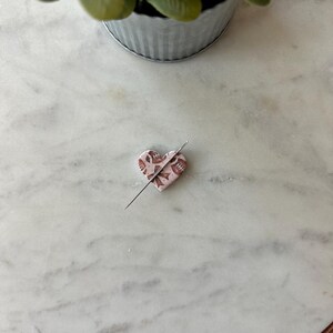 May include: A heart-shaped pin cushion with a floral pattern in shades of pink and brown, pierced by a silver needle. The pin cushion is resting on a white marble surface.